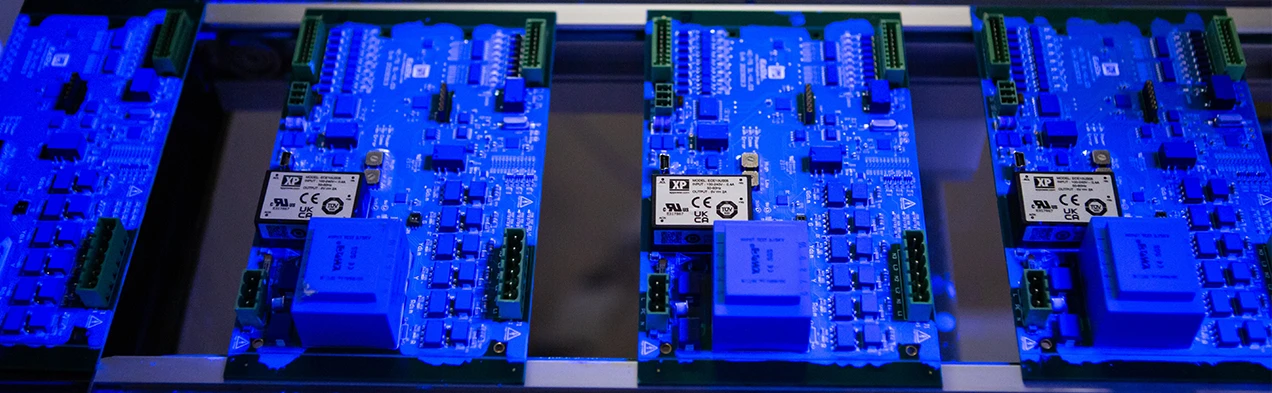 Cette image montre des cartes électroniques passant sous une lumière UV sur un tapis roulant, dans le cadre du processus automatisé de revêtement conforme chez Cloos Electronic. Cette technique protège les composants sensibles et améliore la durabilité du produit.
